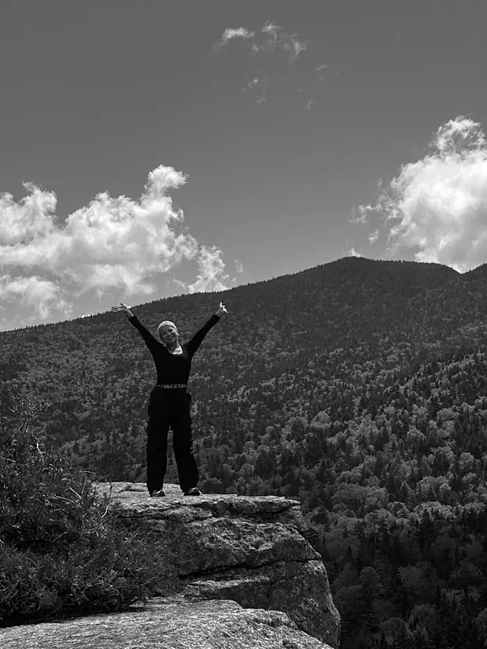 Lauren with arms raised on a rock ledge above a forested valley